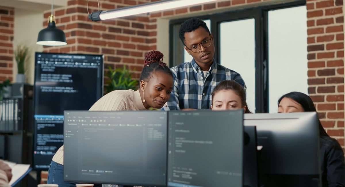 Developers brainstorming ideas looking at code on computer screens asking feedback from senior developer while intern joins the discussion. Junior programmers collaborating on group project.