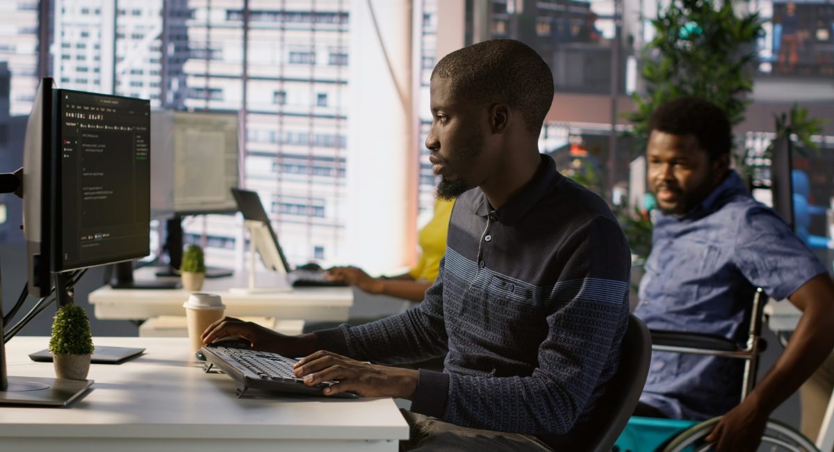 Programmer doing software debugging, asking for help from smiling team leader in wheelchair. Employee fixing coding mistakes, being assisted by cheerful manager with disability, camera A